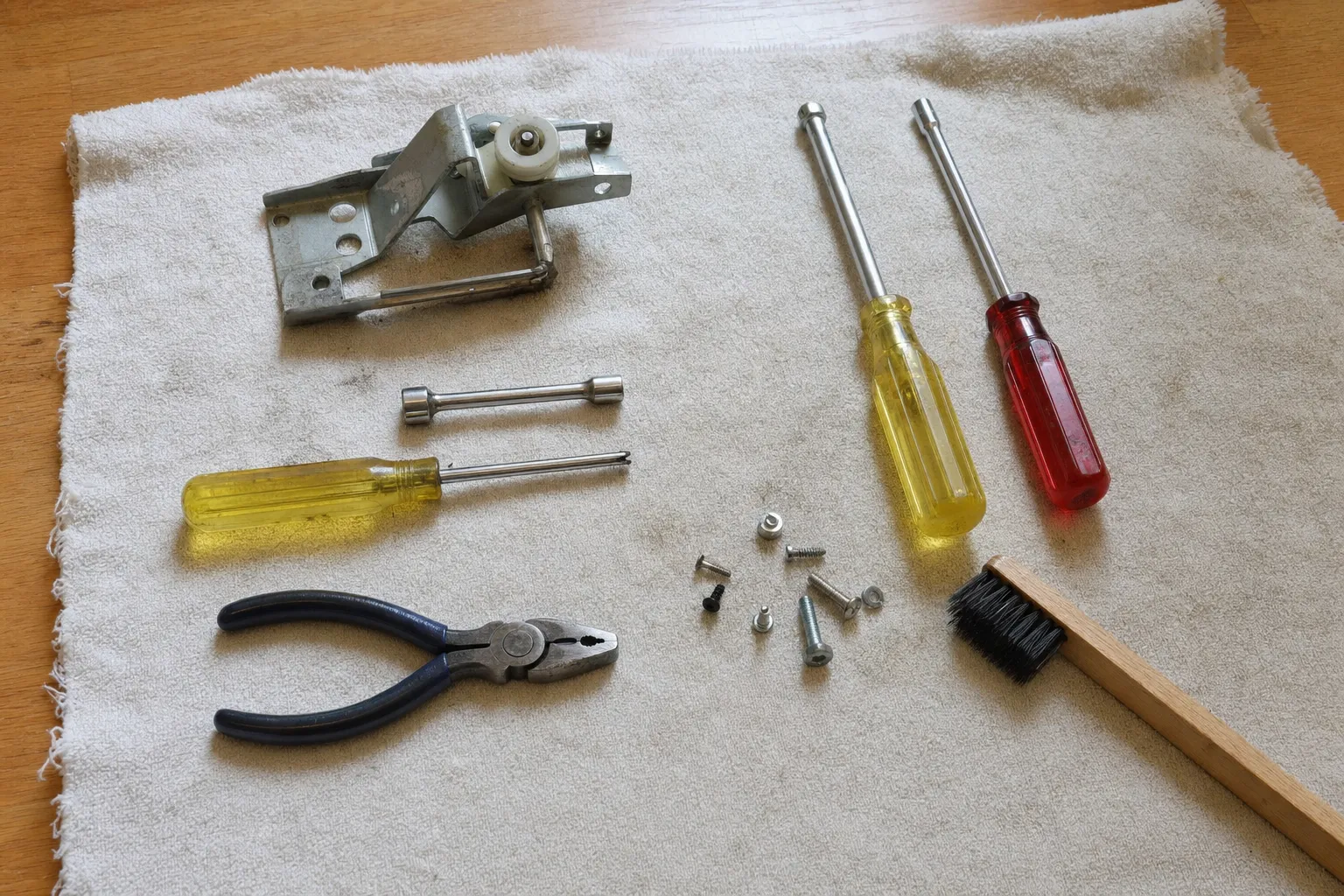 A steel hinge assembly with a white roller, three nut drivers, lineman pliers, loose screws and a wooden brush on a towel